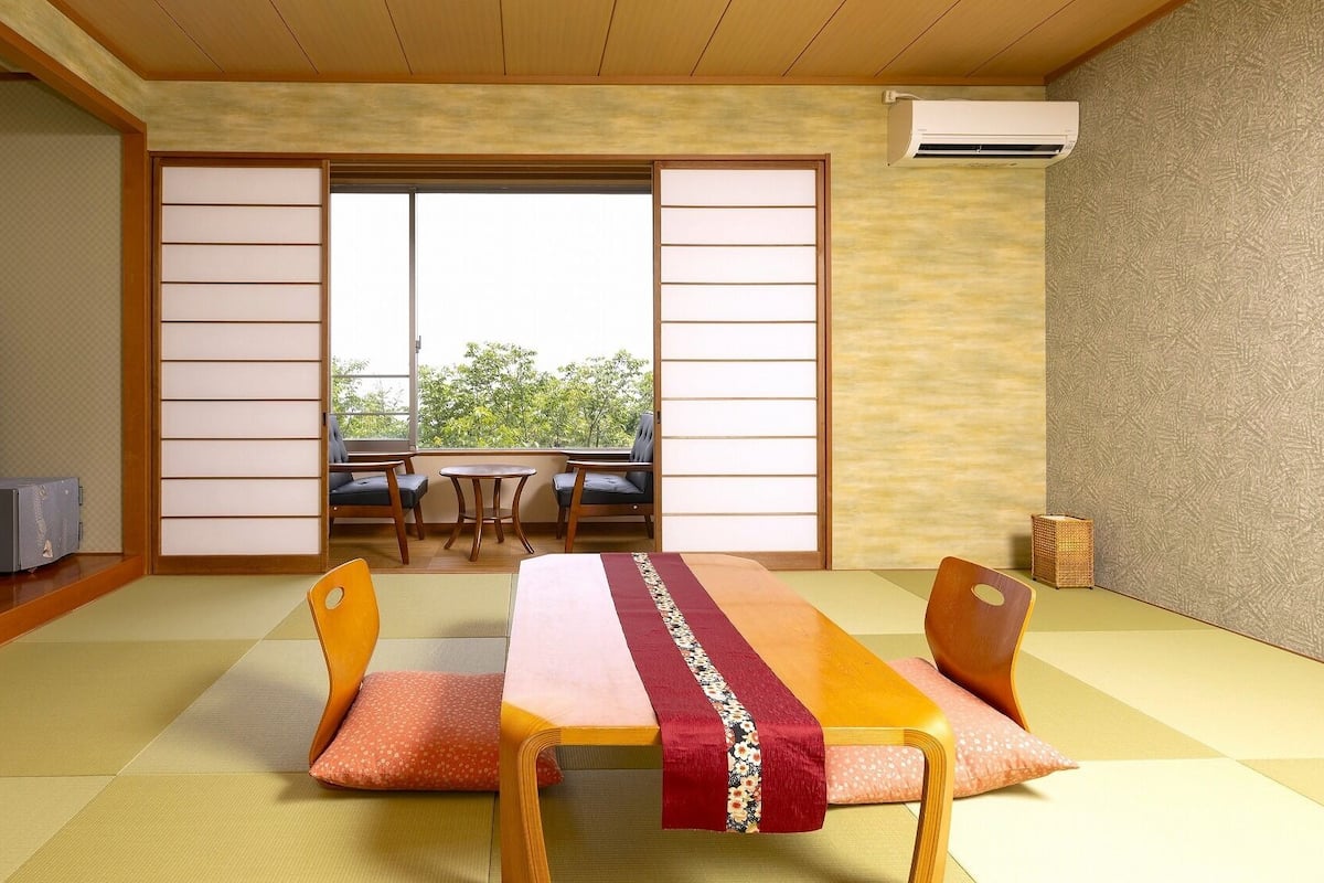 Una habitación con esteras de tatami, una mesa baja, sillas, un televisor y puertas correderas que dan a un balcón.