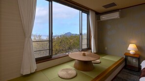 A room with tatami flooring, a low table with a basket, a lamp on a bedside table, and a view of trees and a mountain through large windows.