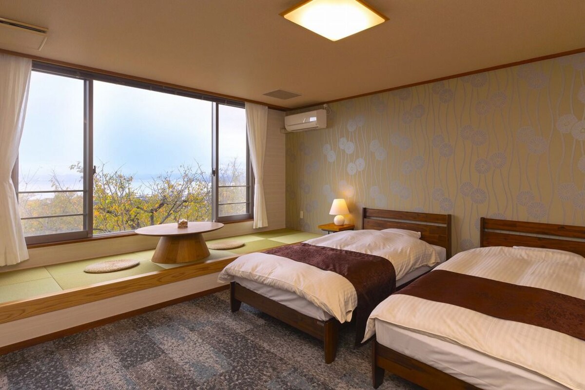 A hotel room with two beds, a window with a view, a bedside table with a lamp, and a small round table with a cushion.