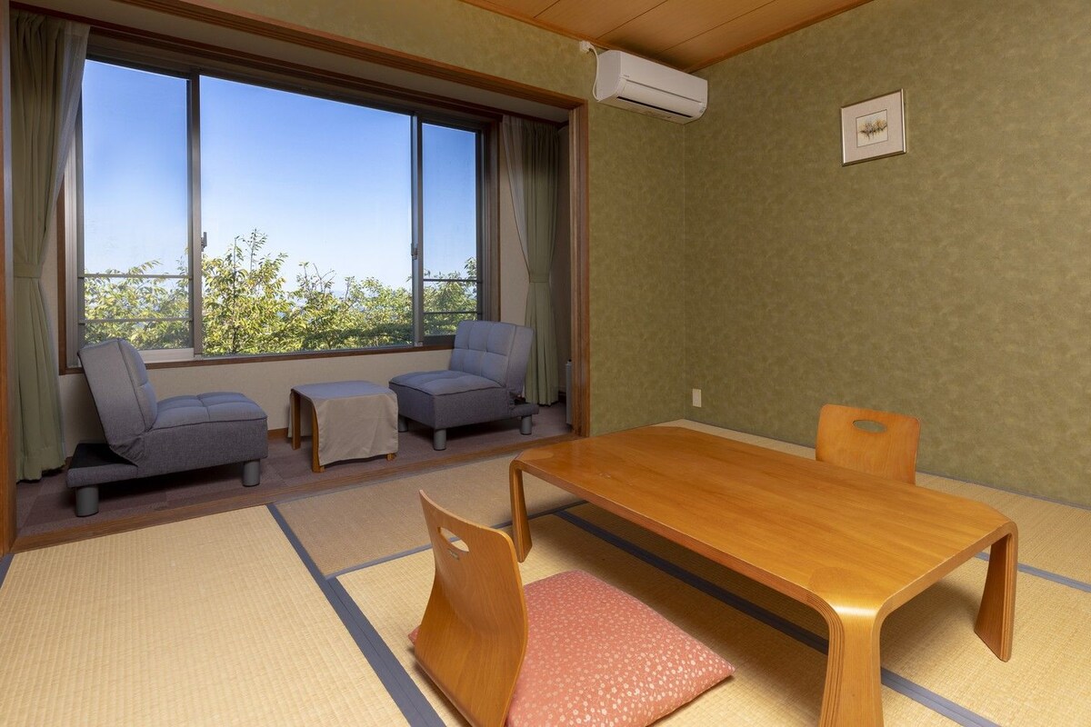 A room with tatami flooring, a wooden table, two grey sofas, and a window with a view of trees.