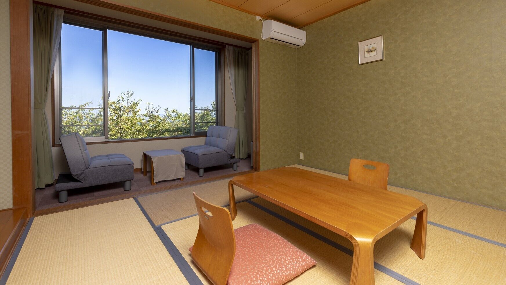 A room with tatami flooring, a wooden table, two grey sofas, and a window with a view of trees.