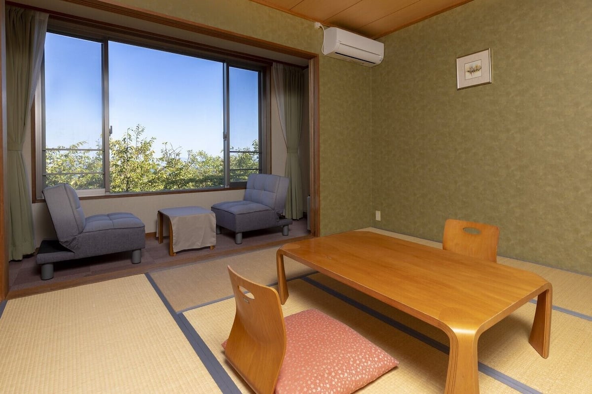 Una habitación con suelo de tatami, una mesa de madera, dos sofás grises y una ventana con vistas a árboles.