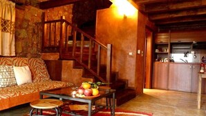 A cozy living room with a wooden staircase, a sofa with patterned cushions, a small round table with fruit, and a kitchen area with wooden cabinets. - Paliokastro Guesthouse (Xylokastro-Evrostina)