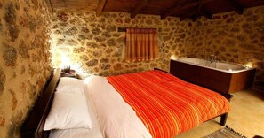 Un dormitorio con pared de piedra, techo de madera, cama con ropa blanca y roja, mesita de noche, mesa auxiliar y un mueble de madera con espejo.