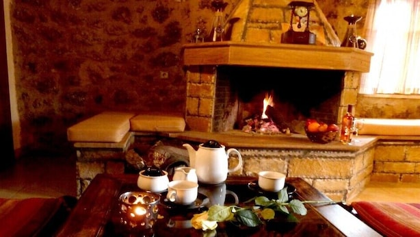 A cozy room with a fireplace, a wooden table with tea set, and a cushioned bench. - Paliokastro Guesthouse (Xylokastro-Evrostina)