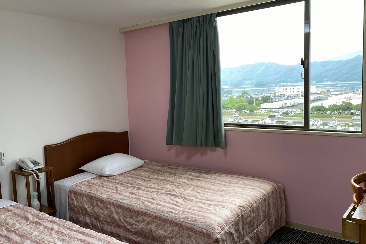 Una camera d'albergo con due letti, un telefono, una finestra vista città e montagne, e una tenda.