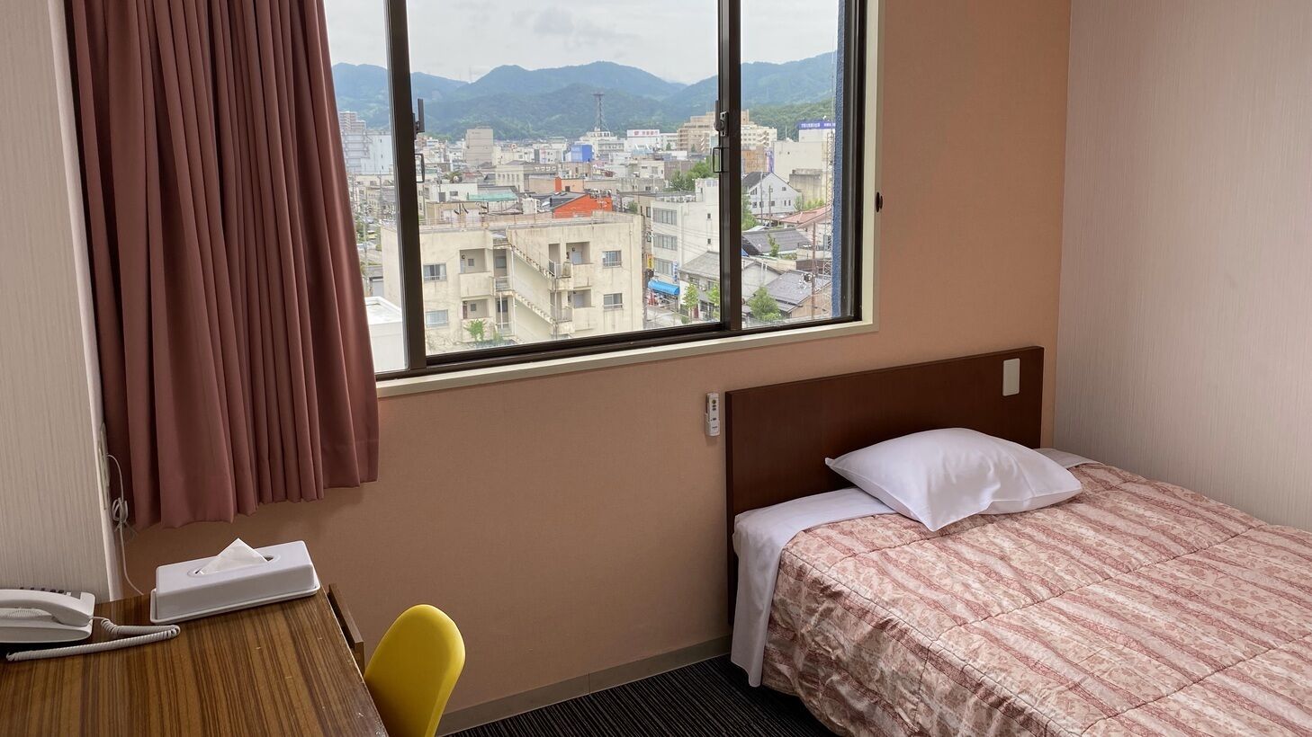 Ein Hotelzimmer mit Bett, Schreibtisch mit Telefon, Stuhl und Fenster mit Blick auf Stadt und Berge.