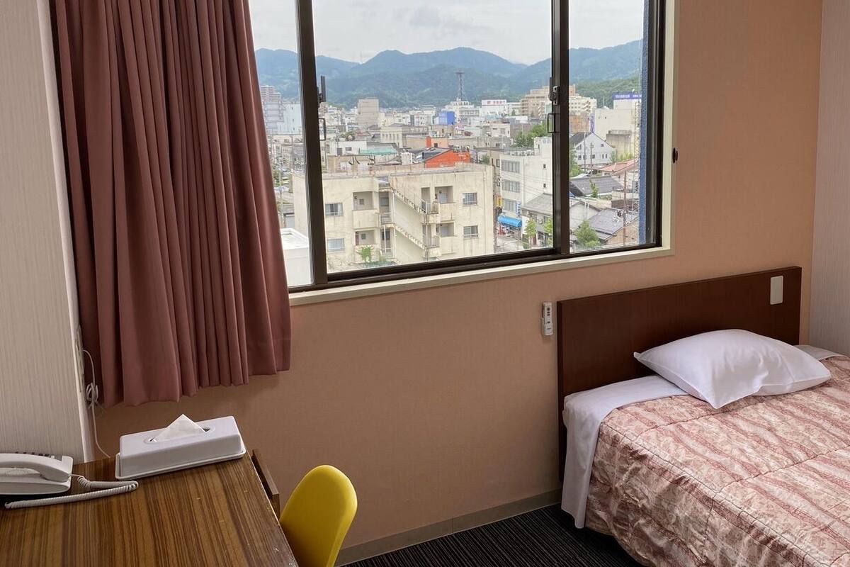 Una camera d'albergo con un letto, una scrivania con il telefono, una sedia e una finestra che si affaccia su una città e montagne.