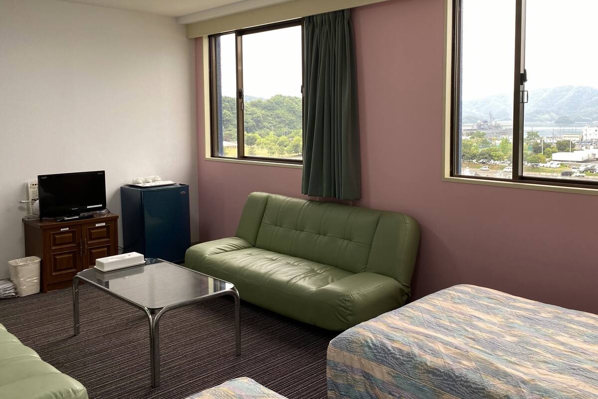 Una camera d'albergo con un divano verde, un letto, una televisione su un mobile e una finestra vista città e montagne.