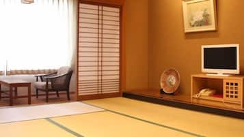 Une pièce de style japonais traditionnel, avec un sol recouvert de tatamis, une petite table avec un coussin, une chaise, un téléviseur posé sur un meuble en bois et une petite étagère sur laquelle se trouvent un téléphone et quelques objets décoratifs.