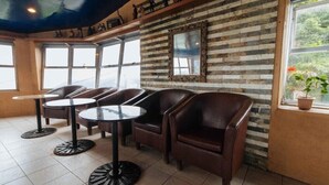 Un café con sillones de cuero y mesas redondas, una pared de ladrillo con un espejo, y ventanas amplias.