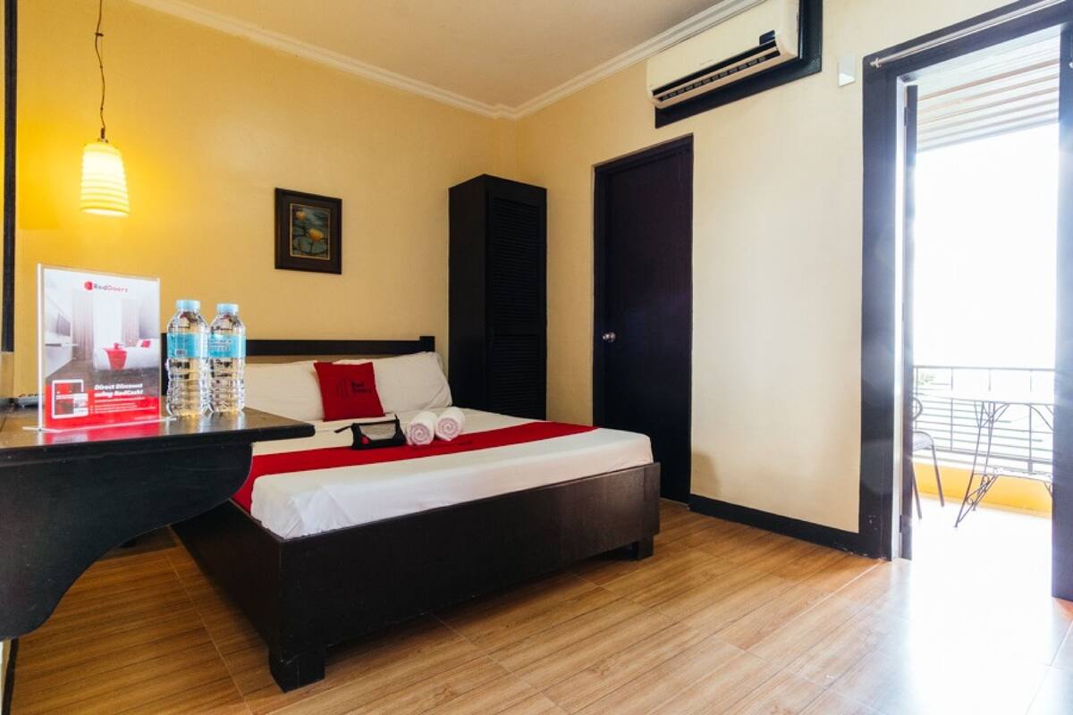 A hotel room with a bed, a desk with water bottles, a red pillow, a black cabinet, and an air conditioning unit.