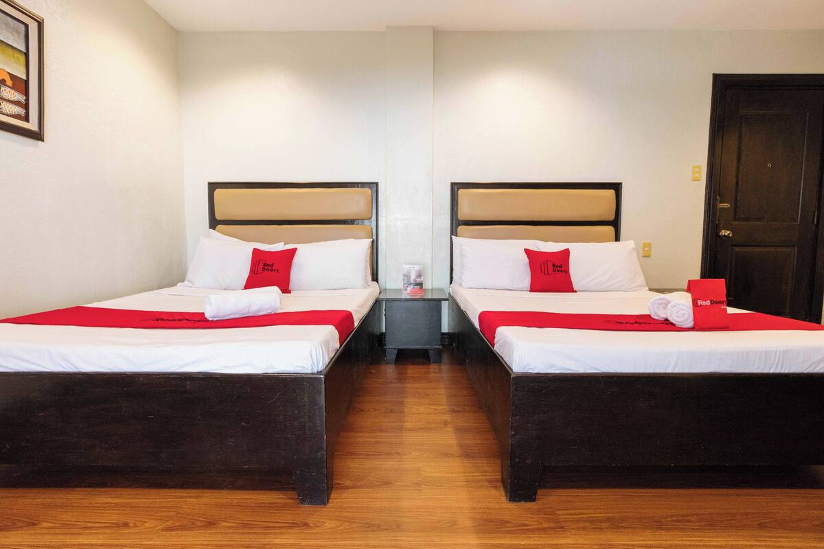A hotel room with two beds, each with a red pillow and a red towel. There is a small table between the beds with a red item on it.
