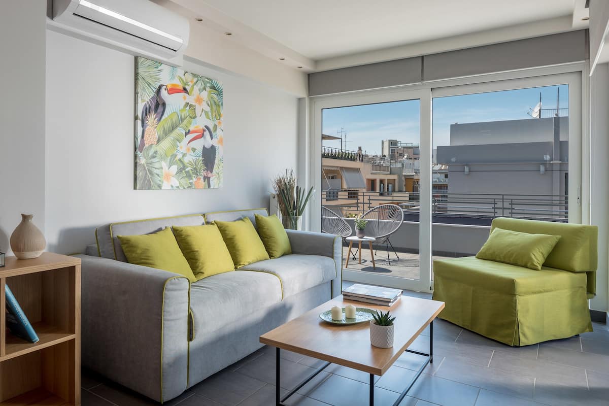 Un salon moderne avec un canapé gris, un fauteuil vert, une table basse en bois et une grande fenêtre donnant sur la ville.