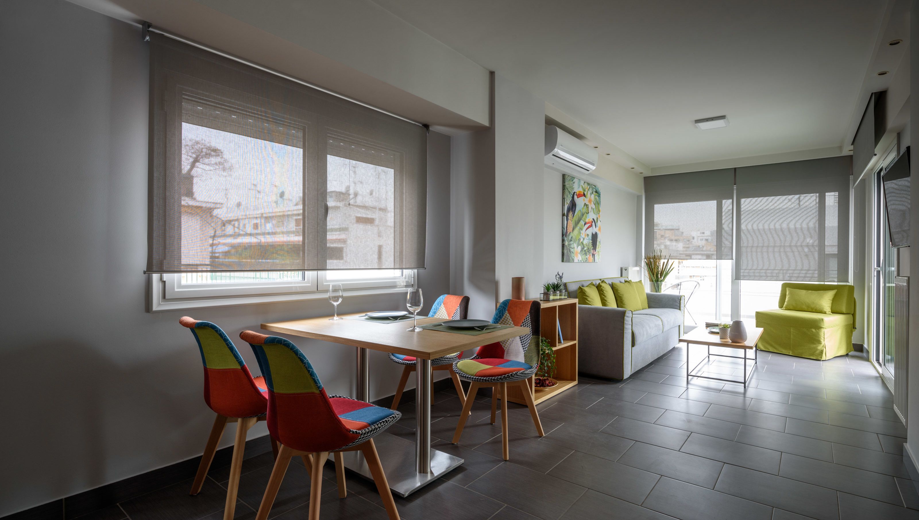 Ein modernes Wohnzimmer mit Essbereich, ausgestattet mit einer Couch, bunten Stühlen, einem Couchtisch und Blick auf die Stadt durch große Fenster.