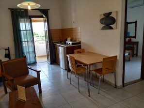 A dining area with a table and chairs, a kitchen with a sink and stove, and a view through a window with curtains. - Pardalakis Studios (Platanias)