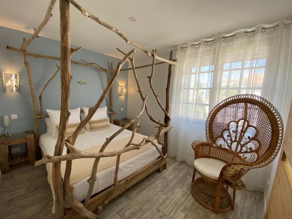 Un dormitorio con cama de dos columnas hecha de ramas, una silla de mimbre y una ventana con cortinas de gasa.