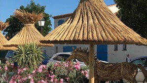 Un edificio con fachada blanca y contraventanas azules, estructura de techo de paja y plantas en floración rosada en primer plano.