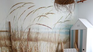 Une pièce comprenant deux chaises en bois, un luminaire suspendu en fibres tressées et une fresque murale représentant de hautes herbes.