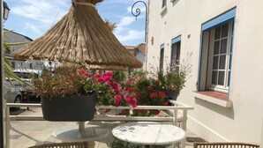 Un área de estar al aire libre con una mesa redonda y sillas de mimbre, un toldo de paja y plantas en macetas con flores rosadas.