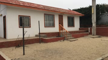 Um edifício térreo com telhado de telhas vermelhas, uma escadaria que leva à entrada e piso de areia.