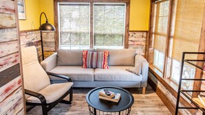 A cozy living room with a sofa, armchair, and coffee table, featuring large windows and wooden walls.