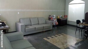 A room with a grey sofa, a small armchair, a desk with a computer, and a bulletin board with papers. - Hotel Castelo (Palmas)