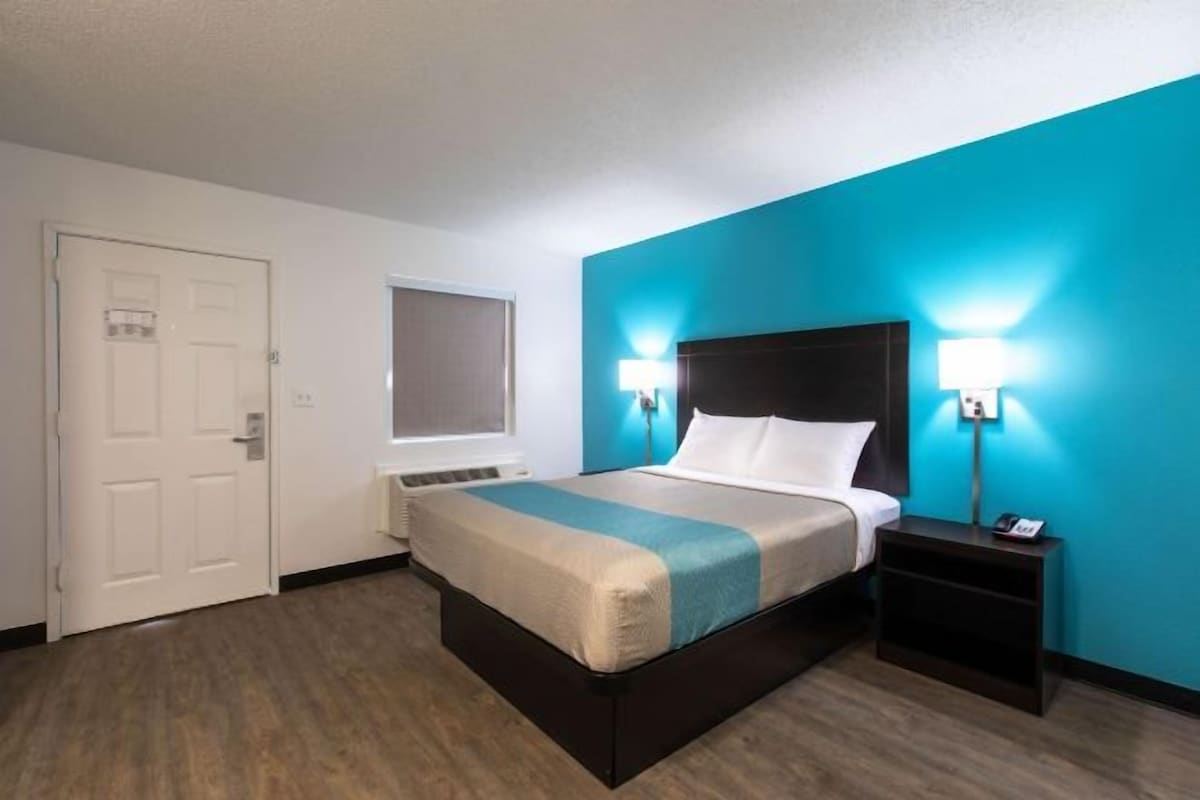 Een hotelkamer met een bed, nachtkastje, lamp en een deur.