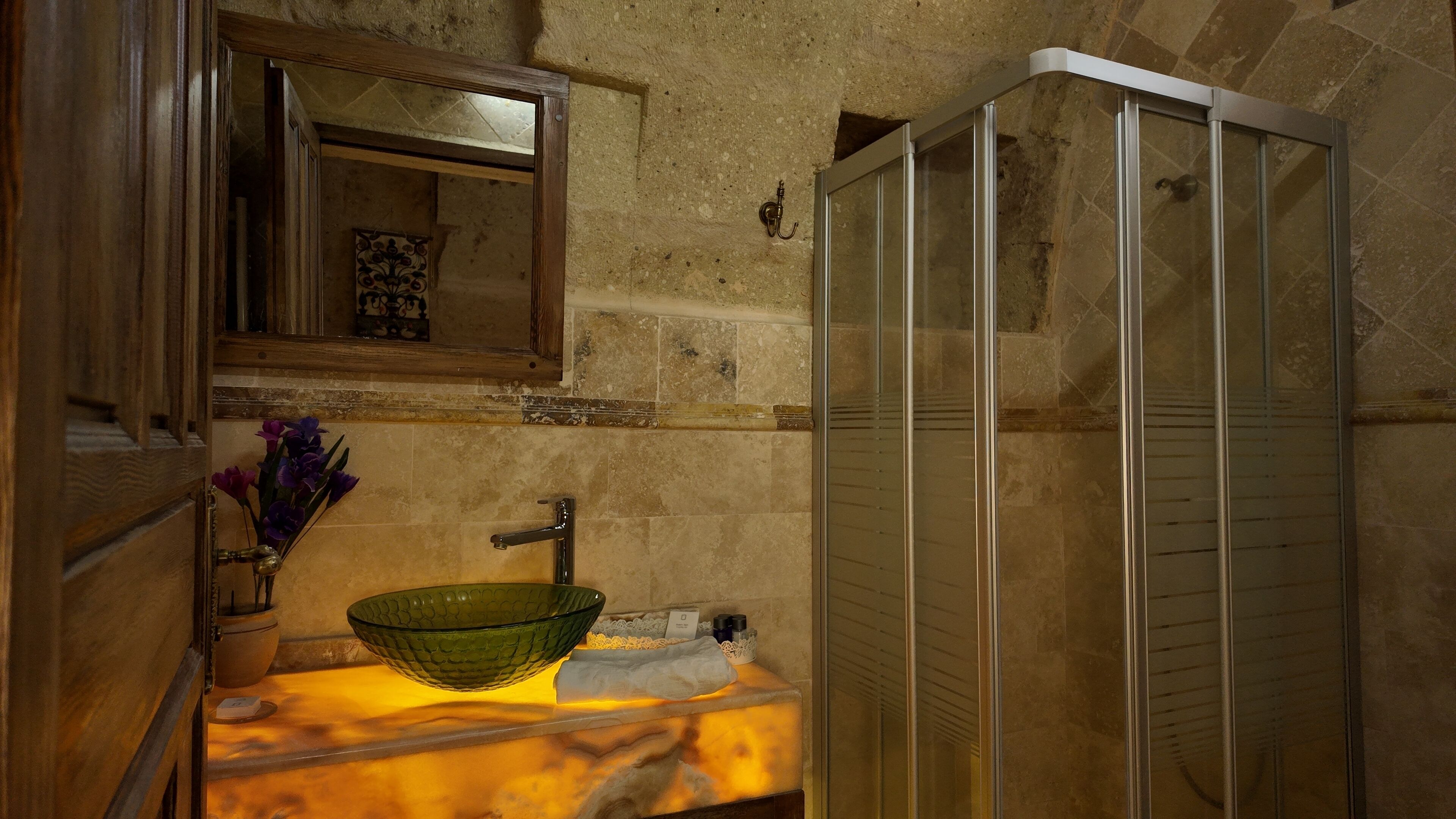 A bathroom with a stone wall, a wooden vanity, a green glass bowl sink, and a glass shower enclosure.