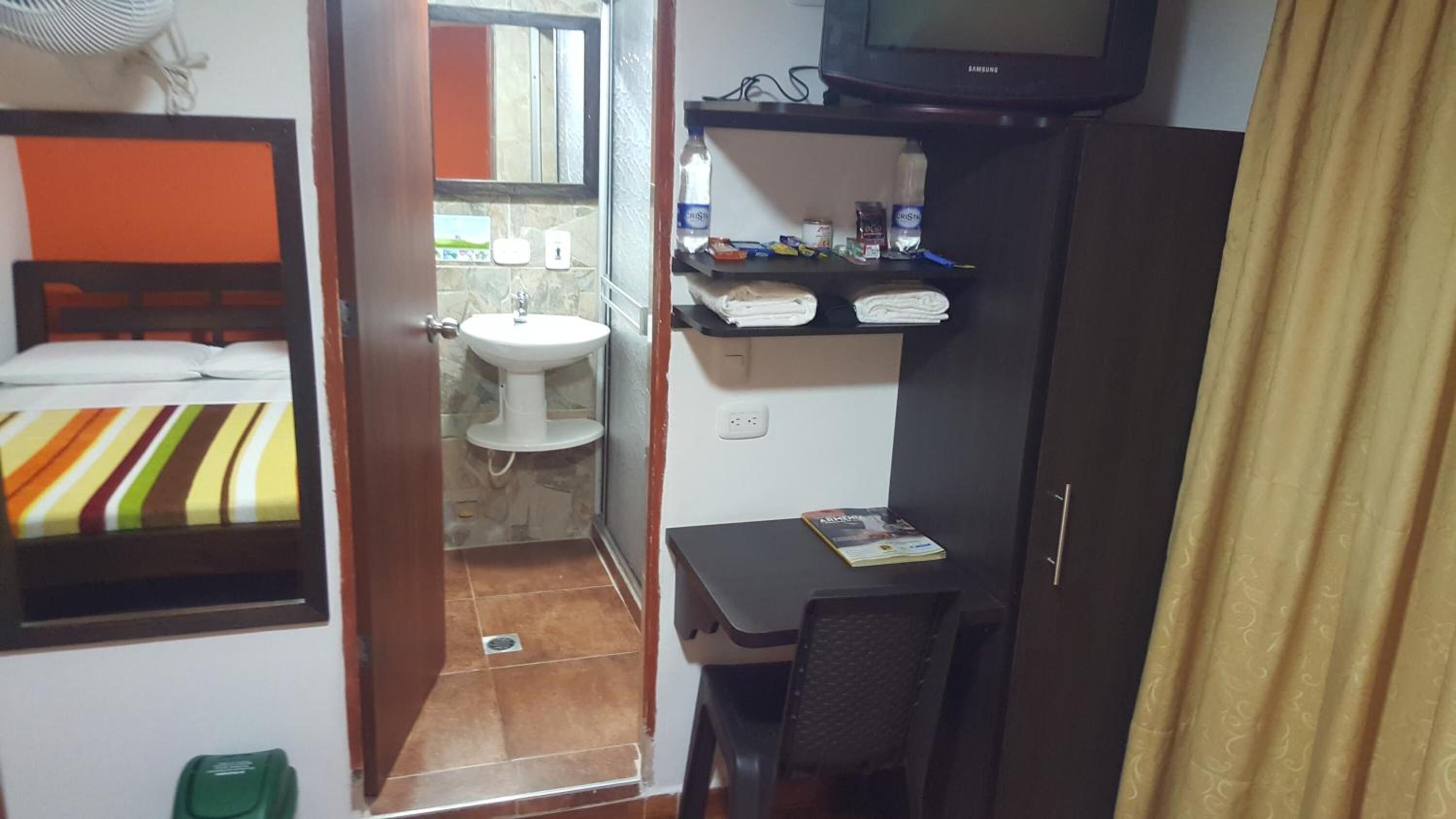 Een kleine hotelkamer met een bed, een bureau, een stoel, een televisie, een plank met toiletartikelen, en een badkamer met een wastafel en spiegel.