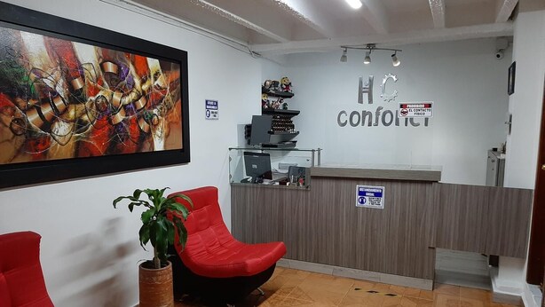 Un área de recepción con una silla roja, una planta en maceta, un cuadro abstracto enmarco y un mostrador de recepción con una computadora y un letrero que dice 