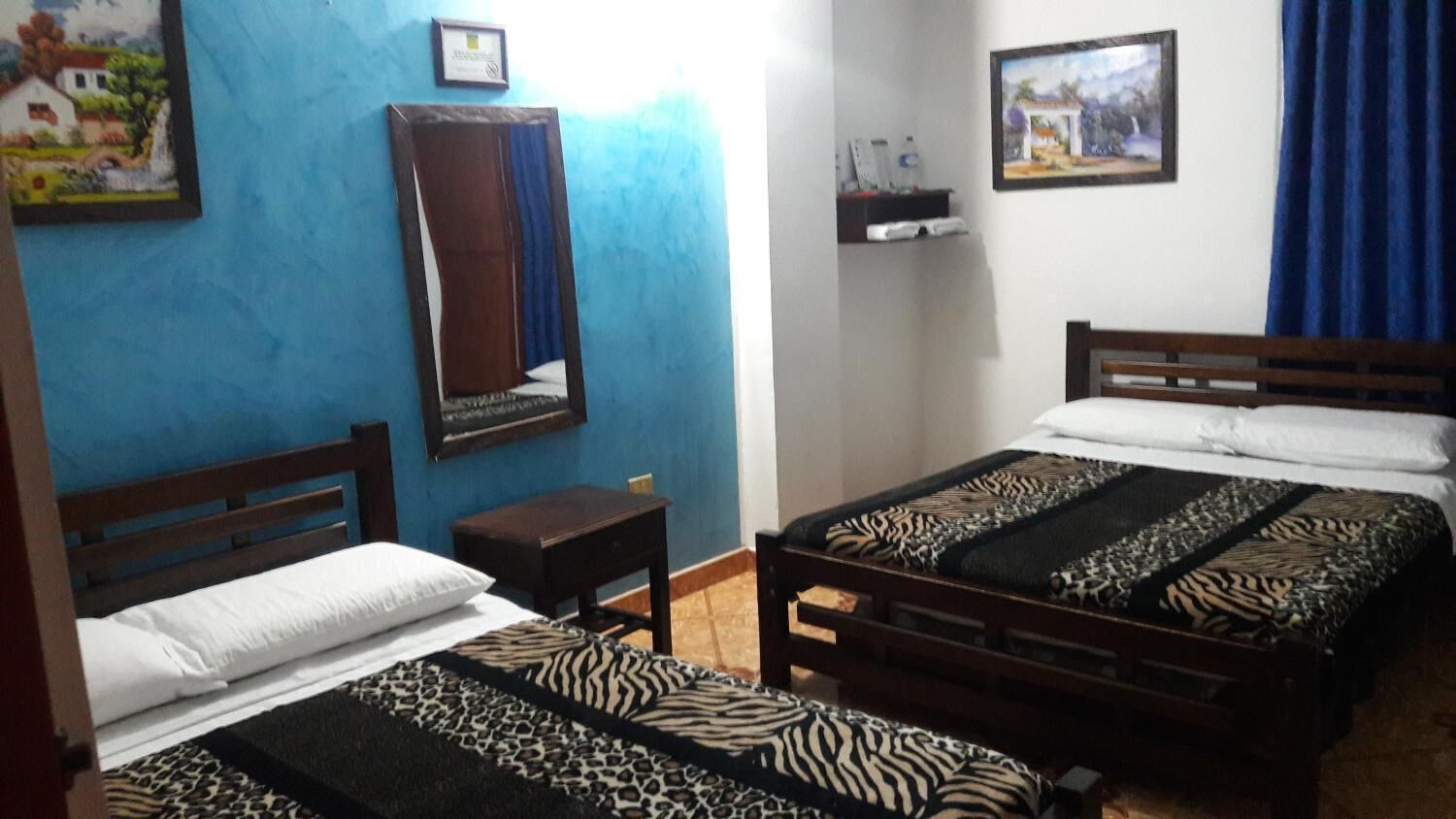 A bedroom with two beds, a small bedside table, a mirror, and two wall-mounted paintings.