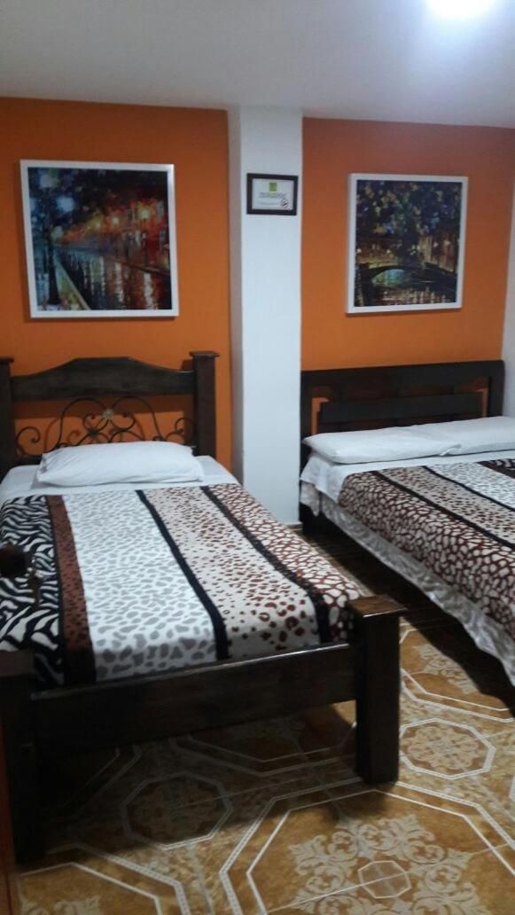 A room with two beds, each with a patterned bedspread and a headboard. The walls are painted orange, and there are two framed pictures on the wall.