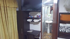 Una habitación pequeña y funcional con cama, mesita de noche, estante con toallas, una pequeña mesa con un libro, televisor montado en la pared, lavamanos y ventilador.