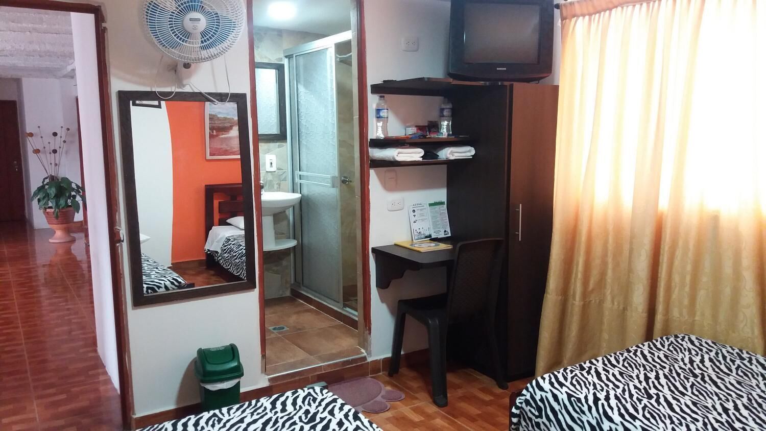 Una habitación compacta con cama, un escritorio pequeño, televisión y baño visible a través de una puerta de cristal.
