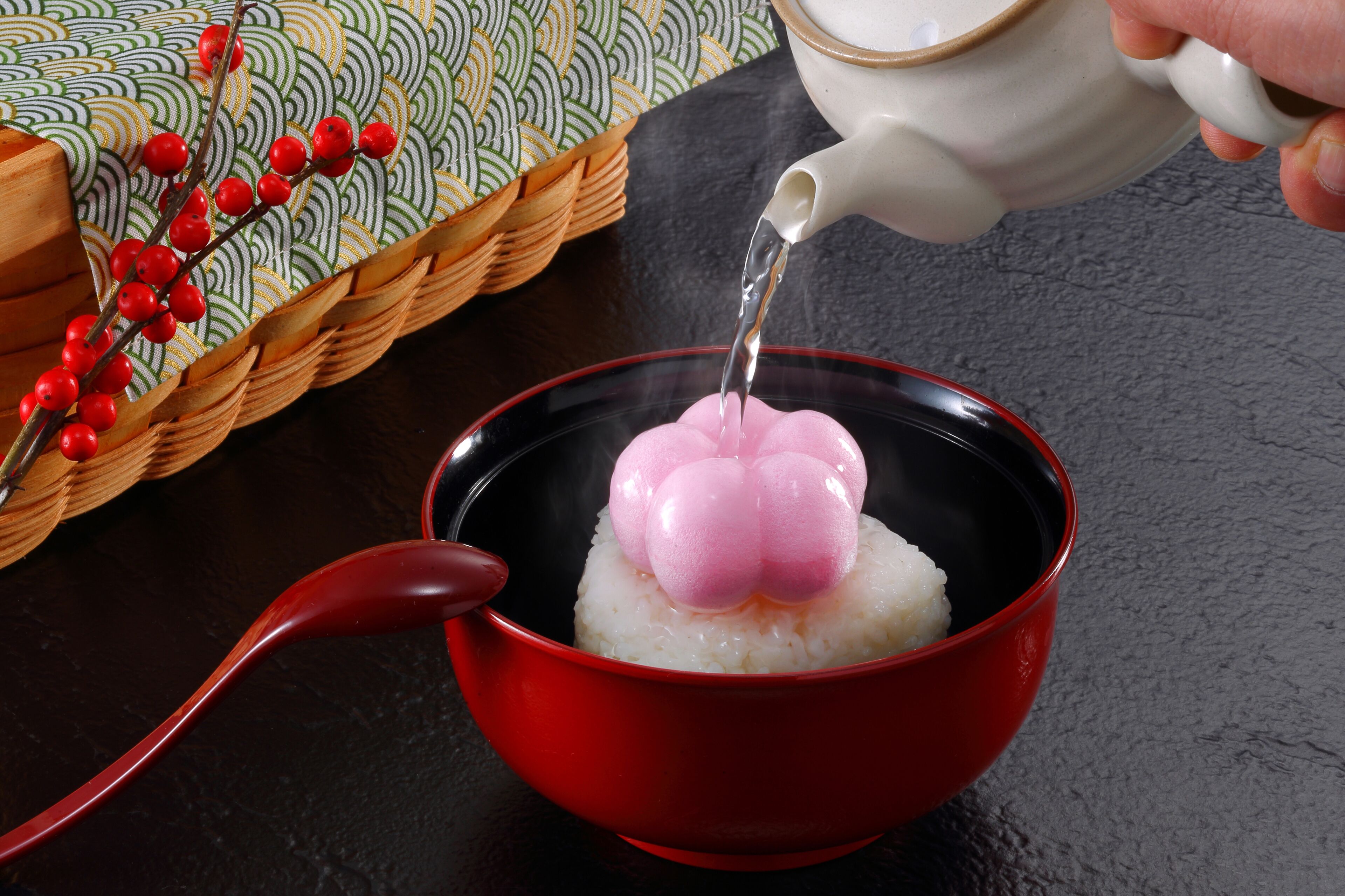 A red bowl with white rice and pink mochi on top, being filled with water from a white teapot.