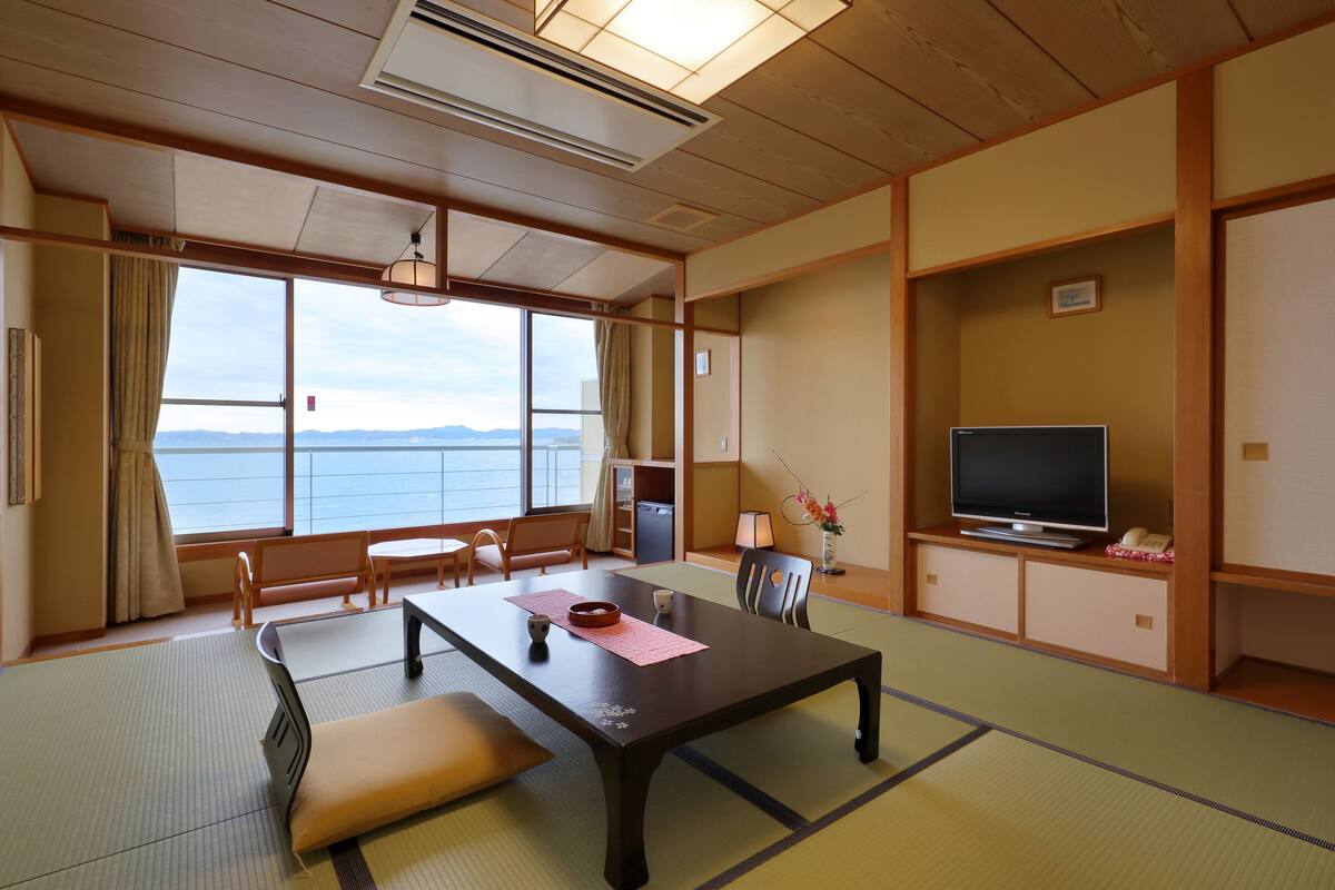 A traditional Japanese room with tatami flooring, a low table with cushions, a television, and a view of the sea.