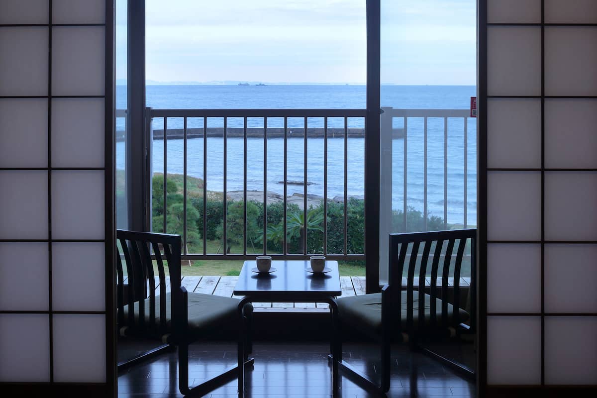 A room with a view of the sea through a large window, a wooden table with two chairs, and a traditional Japanese shoji screen.