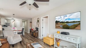 Ein geräumiges Wohnzimmer mit Deckenventilator, einem großen Flachbildfernseher an der Wand, einem gelben Hocker und einem Couchtisch aus Holz.