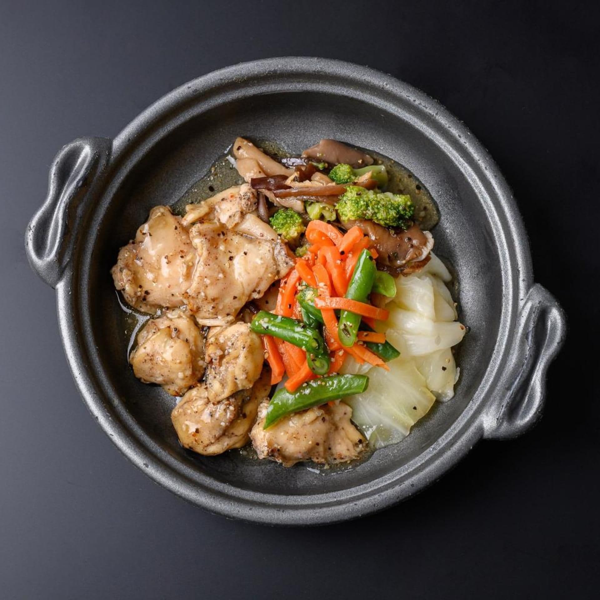 Panela redonda escura com alças, contendo um refogado de frango, legumes e molho.