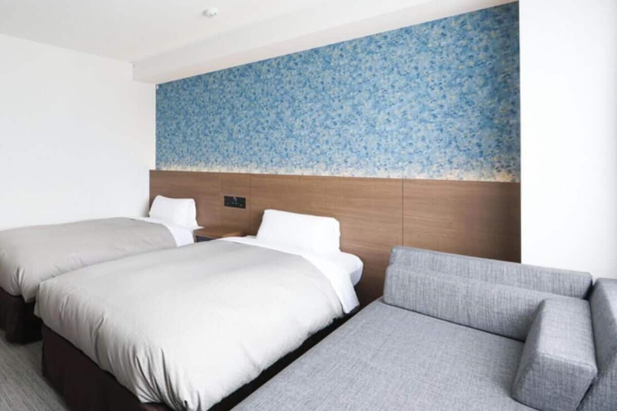 A hotel room with two beds, a grey sofa, and a blue floral wall.