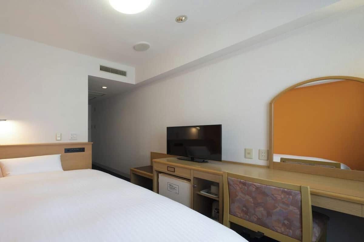 A hotel room with a bed, a desk with a television, a small refrigerator, and a mirror.