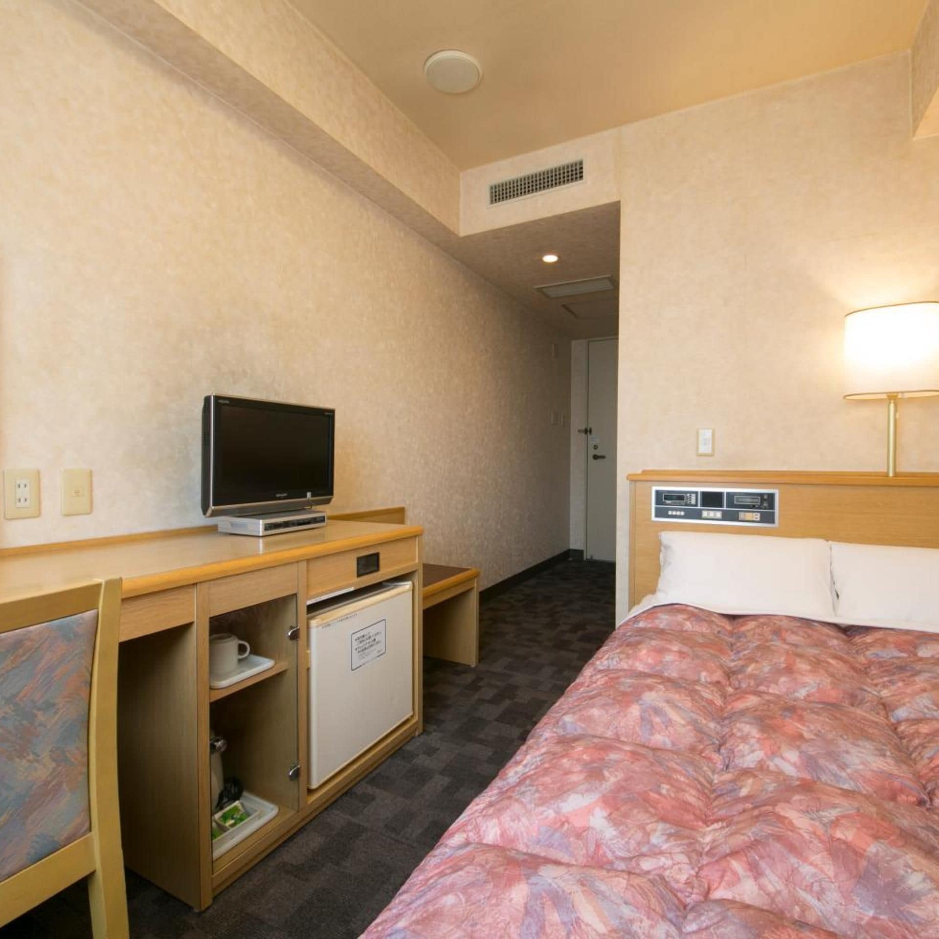 A hotel room with a bed, a desk with a television, a mini-fridge, and a lamp.