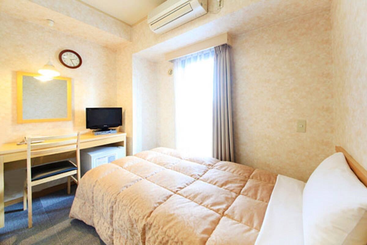 A hotel room with a bed, a desk with a television, a clock, and an air conditioning unit.