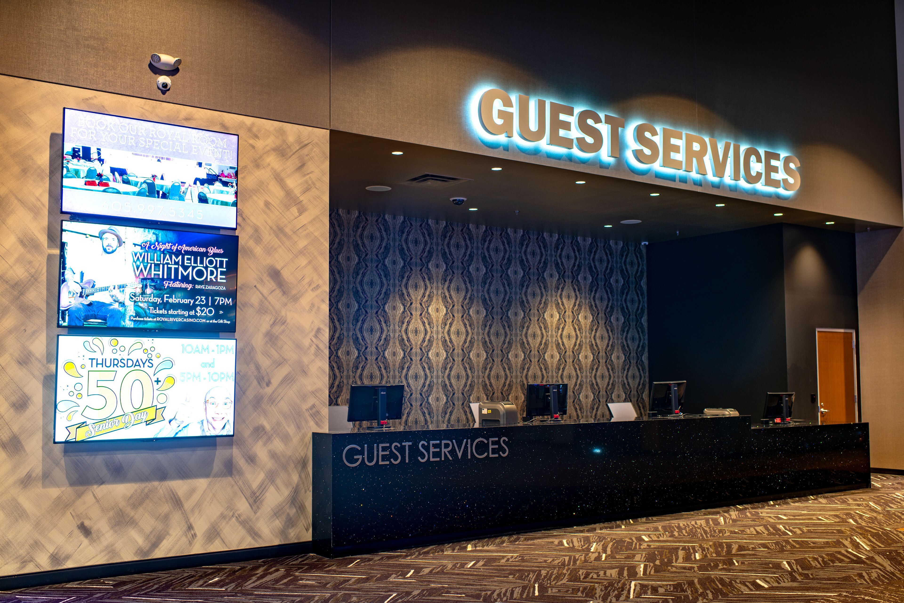 A hotel reception area with a 'Guest Services' sign, a patterned wall, and a digital screen displaying event information.
