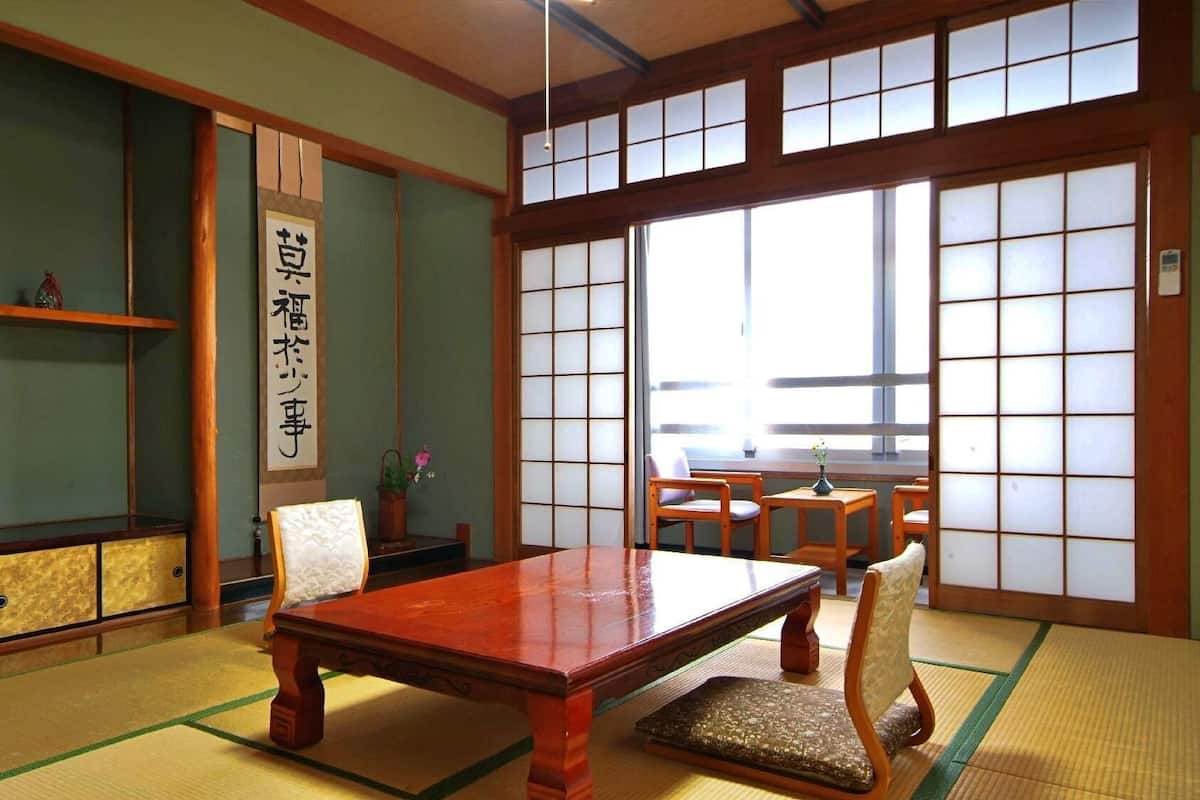 Ein traditioneller japanischer Raum mit Tatami-Boden, einem niedrigen Tisch, Stühlen, einem Fernseher und verschiebbaren Türen.
