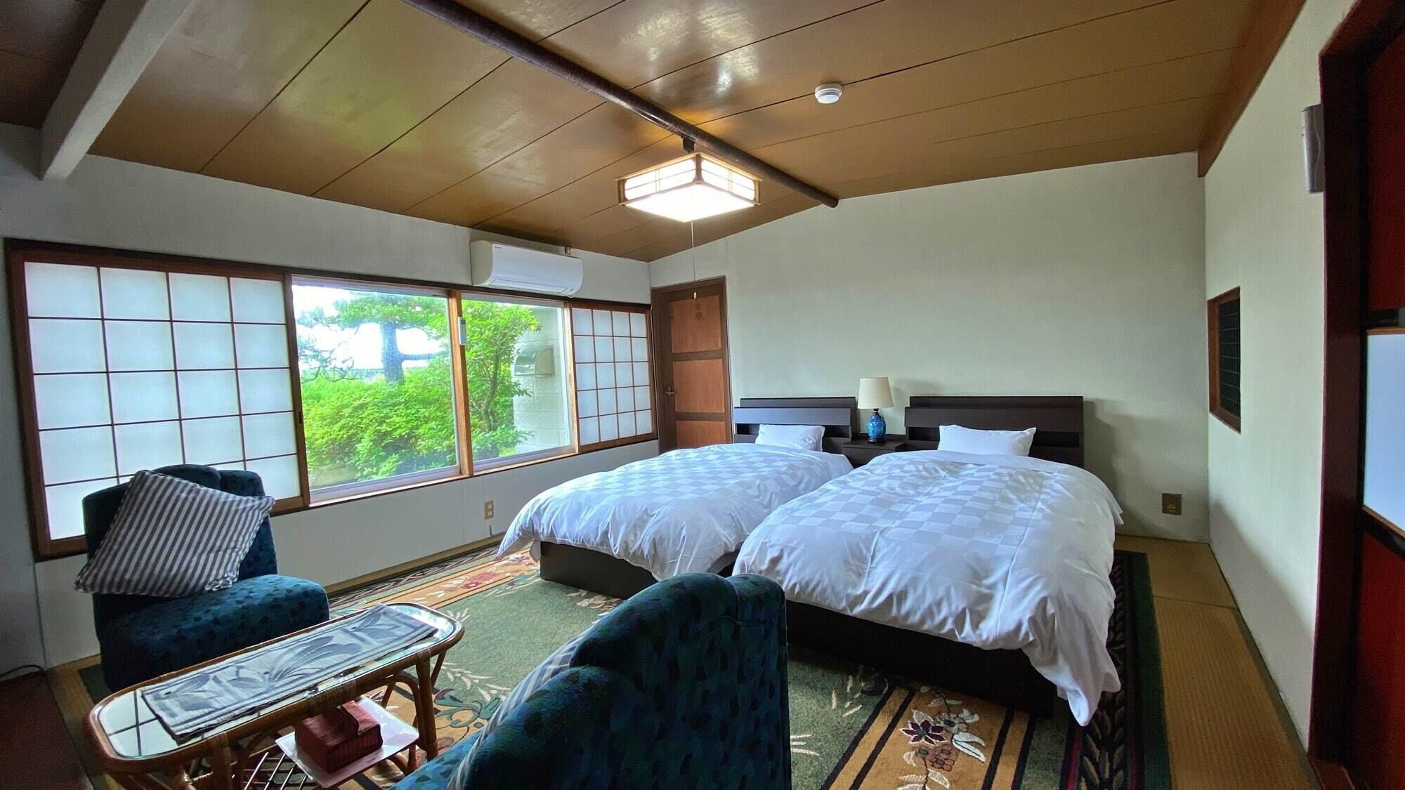 A hotel room with two beds, a sitting area with a chair and table, large windows with a view, and a ceiling fan.