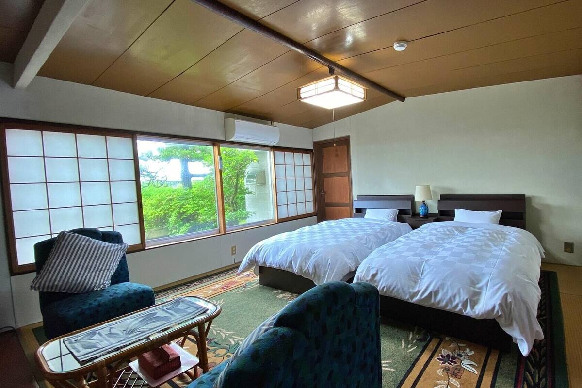 Ein Hotelzimmer mit zwei Betten, einer Sitzecke mit Stuhl und Tisch, großen Fenstern mit Aussicht und einem Deckenventilator.
