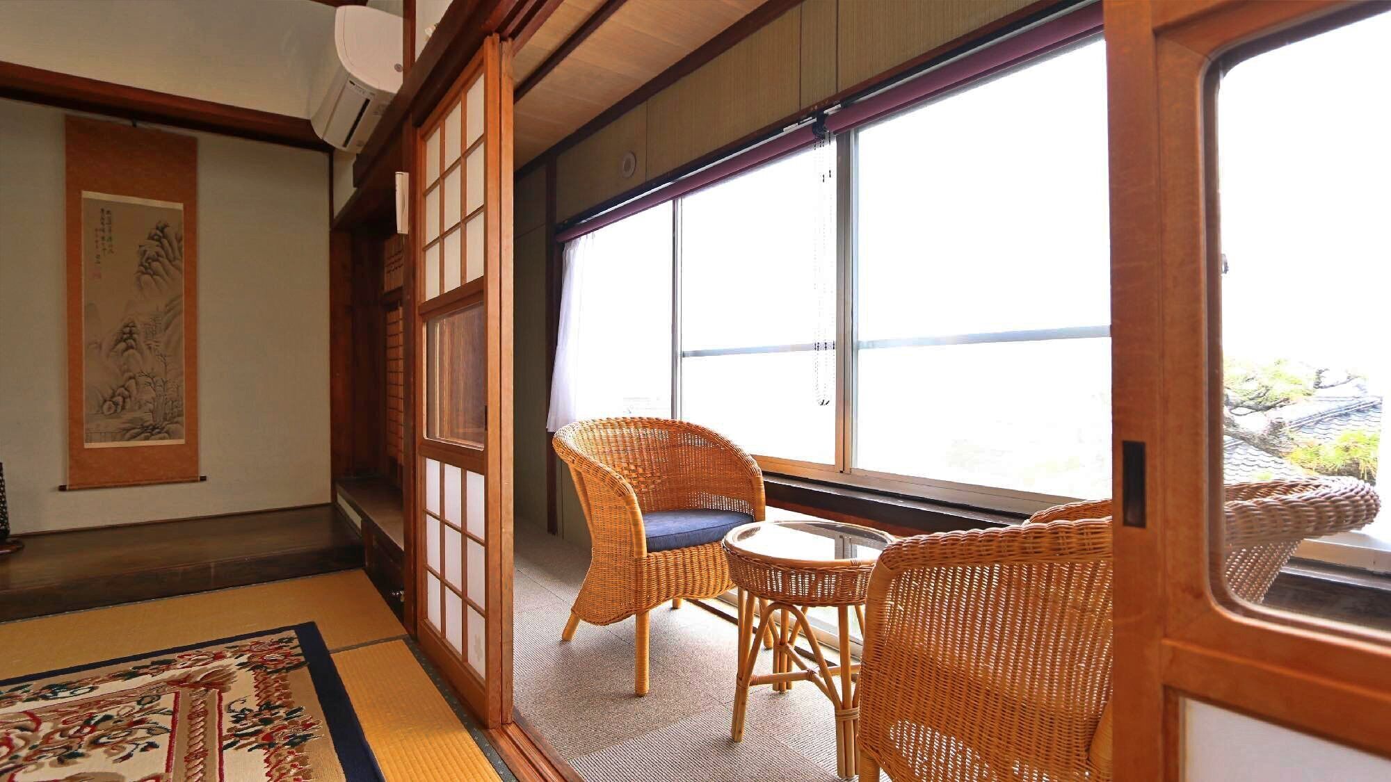A traditional Japanese room with tatami flooring, a low table with two wicker chairs, and a large window with a view.