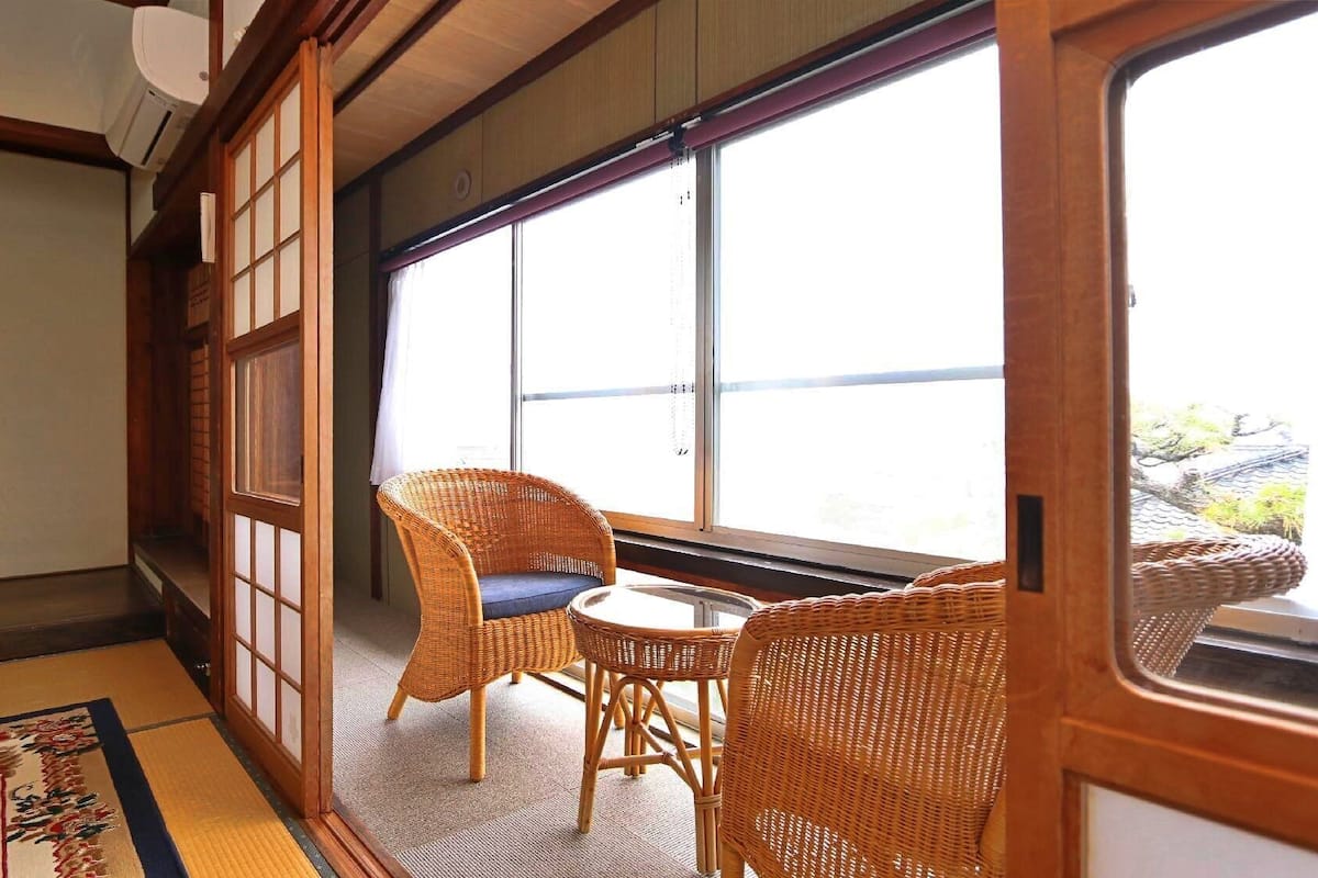 Ein traditioneller japanischer Raum mit Tatami-Böden, einem niedrigen Tisch mit zwei Korbstühlen und einem großen Fenster mit Aussicht.
